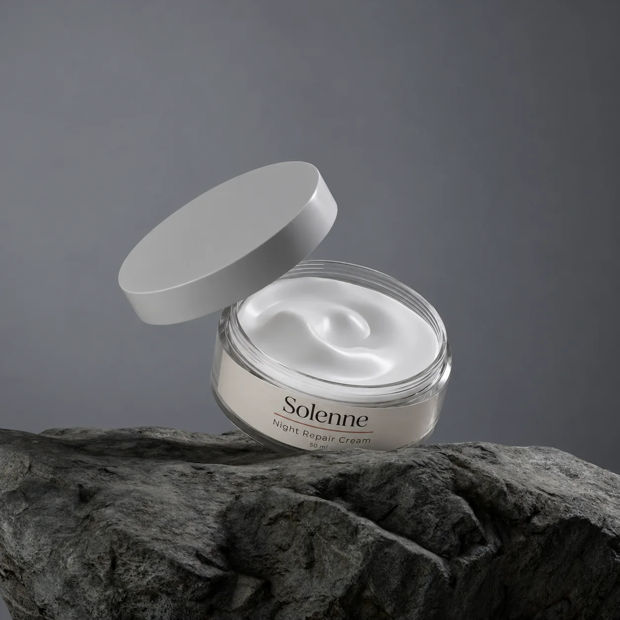 An open jar of Night Repair Cream, its lid tipped back, on a dark rock